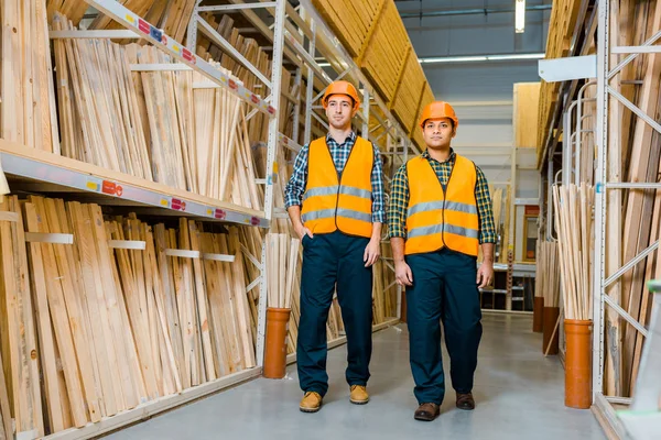 Multikulti-Lagerarbeiter in Schutzanzügen und Helmen gehen an Regalen mit Holzbaumaterialien entlang — Stockfoto