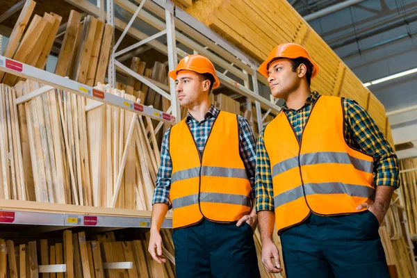 Multicultural warehouse workers in safety vasts and helmets standing near racks with wooden construction materials — Stock Photo