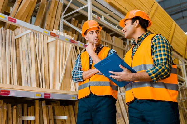 Trabajadores multiculturales reflexivos de pie cerca de estantes con materiales de construcción de madera - foto de stock