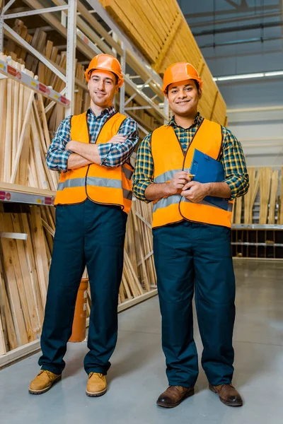 Trabalhadores multiculturais sorridentes em coletes de segurança e capacetes sorrindo e olhando para a câmera — Fotografia de Stock