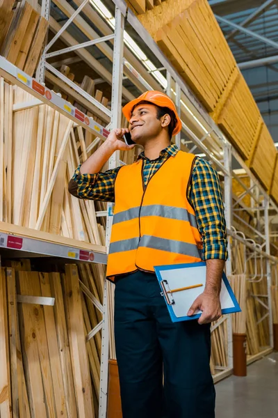 Alegre trabajador de almacén hablando en el teléfono inteligente y sujetando portapapeles - foto de stock