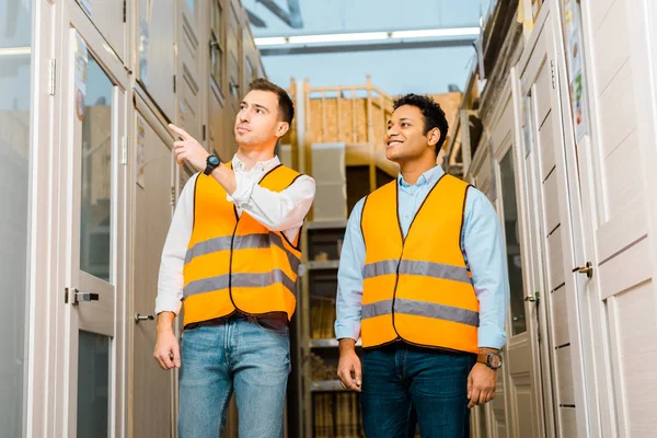 Guapo trabajador de almacén señalando con el dedo a las puertas cerca sonriente indio compañero de trabajo - foto de stock