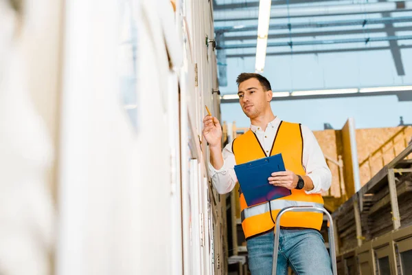 Selektiver Fokus eines gut aussehenden, aufmerksamen Arbeiters, der auf einer Leiter steht und Klemmbrett hält — Stockfoto