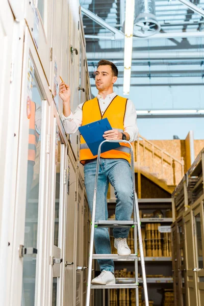 Enfoque selectivo del trabajador concentrado en chaleco de seguridad de pie en la escalera en el departamento de puertas - foto de stock