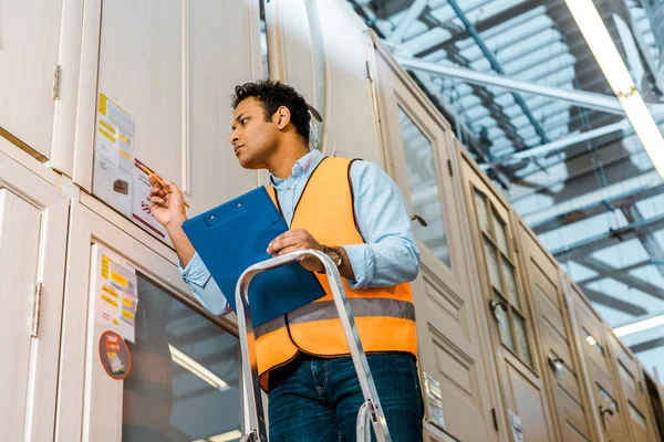 Gutaussehender indischer Arbeiter in Sicherheitsweste steht auf Leiter im Lager — Stockfoto