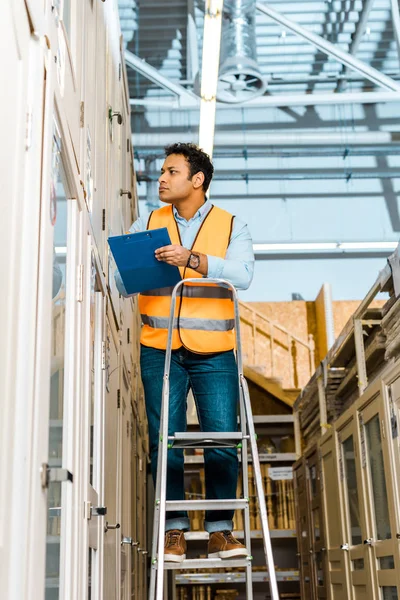 Selective focus of attentive indian worker in safety vest standing on ladder in warehouse — Stock Photo