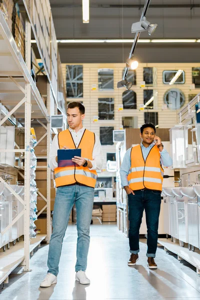 Schöner indischer Arbeiter spricht auf Smartphone neben Kollege mit Klemmbrett — Stockfoto