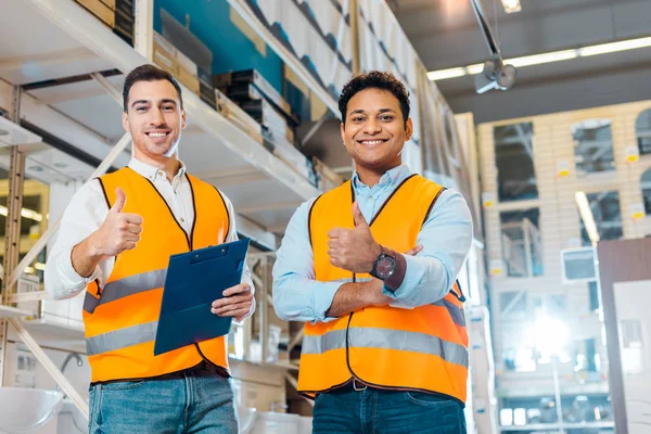 Alegres trabajadores de almacenes multiculturales mostrando pulgares hacia arriba y mirando a la cámara - foto de stock