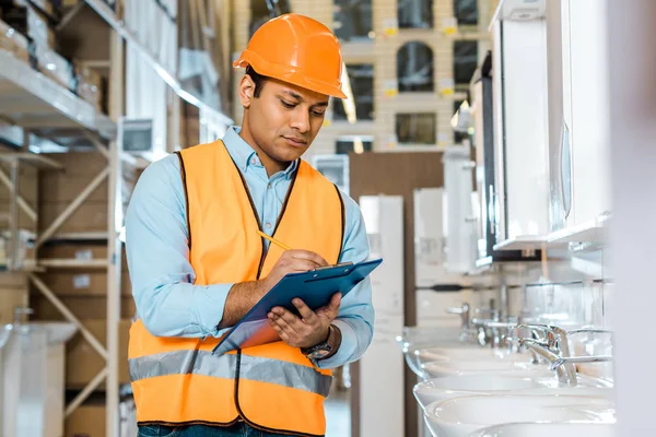 Selective focus of serious indian worker writing in clipboard in plumbing department — Stock Photo