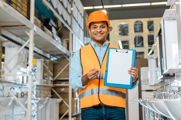 Smiling indian warehouse worker showing clipboard with blank paper and looking at camera — Stock Photo