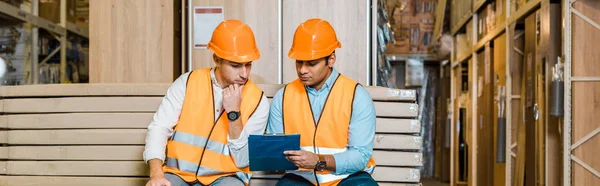 Panoramic shot of serious multicultural workers sitting on bench in warehouse and looking at clipboard — Stock Photo