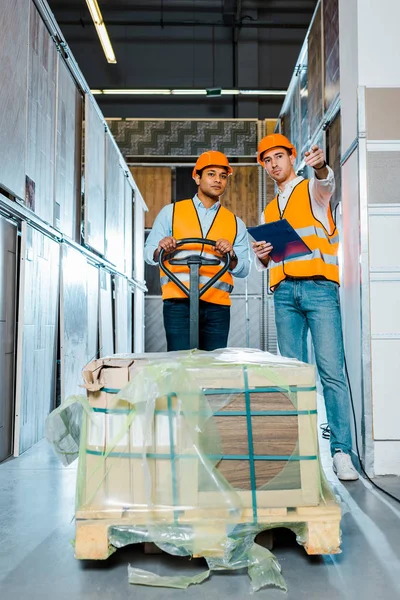 Multicultural warehouse workers in helmets and safety vasts standing near pallet jack in tiles department — Stock Photo