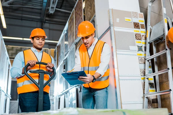 Upset indian worker with pallet jack standing near angry colleague — Stock Photo