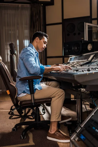 Productor de sonido de carrera mixta atento trabajando en la consola de mezcla en el estudio de grabación - foto de stock