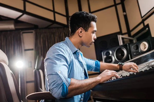 Apuesto músico de carrera mixta trabajando en la consola de mezcla en el estudio de grabación - foto de stock