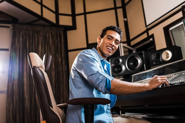 Fröhlicher Mixed Race Soundproduzent sitzt im Bürostuhl im Tonstudio und blickt in die Kamera — Stockfoto