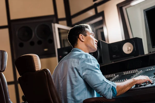 Productor de sonido de carrera mixta atento trabajando en la consola de mezcla en el estudio de grabación - foto de stock