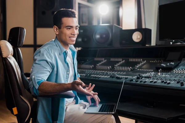 Smiling mixed race sound producer sitting by mixing console and using laptop — Stock Photo