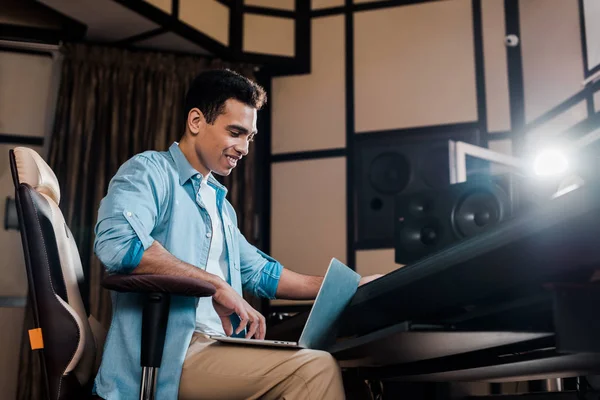 Smiling mixed race sound producer sitting in recording studio and using laptop — Stock Photo