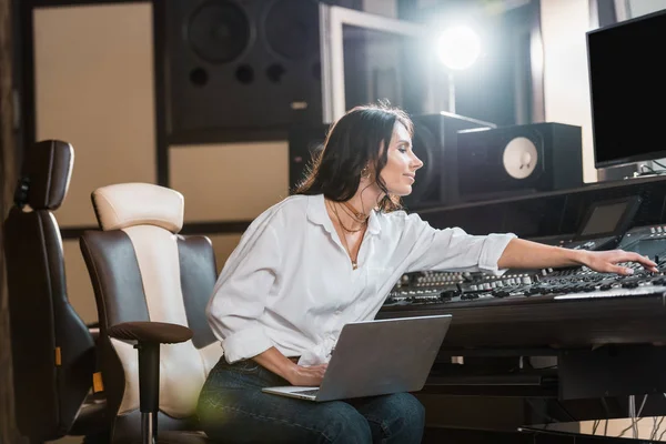 Привлекательный, улыбающийся звукорежиссер, использующий ноутбук и работающий на микширующей консоли в студии звукозаписи — стоковое фото