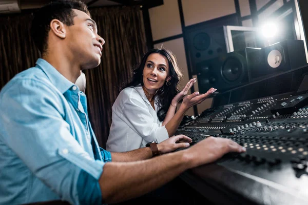 Enfoque selectivo de productor de sonido bastante gesto cerca de compañero de carrera mixta trabajando en la consola de mezcla - foto de stock