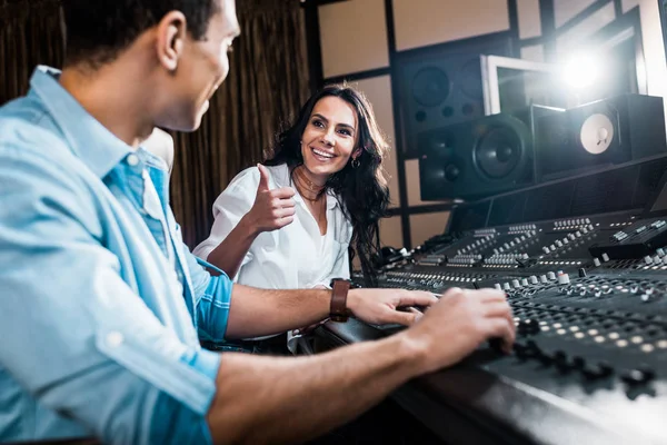 Enfoque selectivo de productor de sonido bastante mostrando el pulgar hacia arriba cerca apuesto colega de raza mixta que trabaja en la consola de mezcla - foto de stock