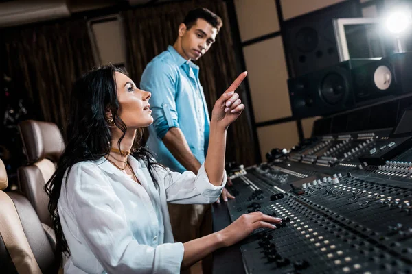 Hübscher Soundproduzent, der mit dem Finger zeigt, während er im Aufnahmestudio in der Nähe eines Mixed Race Kollegen arbeitet — Stockfoto