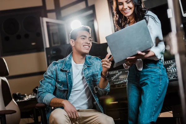 Productores sonrientes de sonido multicultural utilizando el ordenador portátil en el estudio de grabación - foto de stock