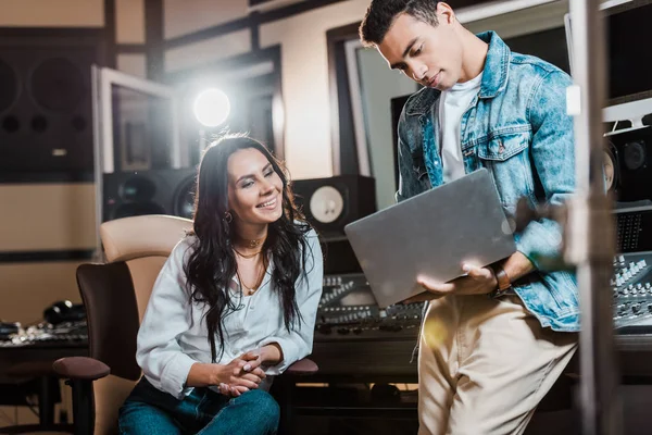 Schöner Mixed Race Soundproduzent mit Laptop in der Nähe eines fröhlich lächelnden Kollegen — Stockfoto