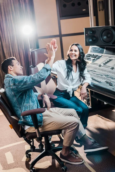 Dos productores de sonido multicultural alegre dando cinco en el estudio de grabación mientras se sienta cerca de la consola de mezcla - foto de stock