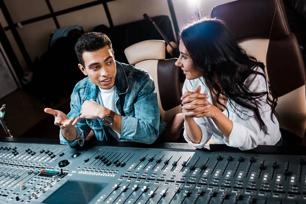 Deux producteurs sonores multiculturels souriants parlent alors qu'ils sont assis près de la console de mixage dans un studio d'enregistrement — Photo de stock