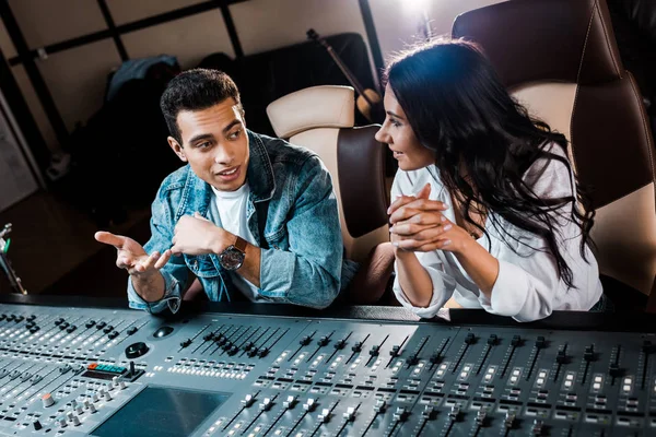 Two smiling multicultural sound producers talking while sitting near mixing console in recording studio — Stock Photo
