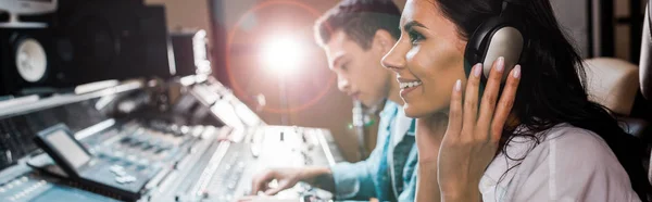 Panoramic shot of attractive sound producer in headphones near mixed racial friend working at mixing console — Stock Photo