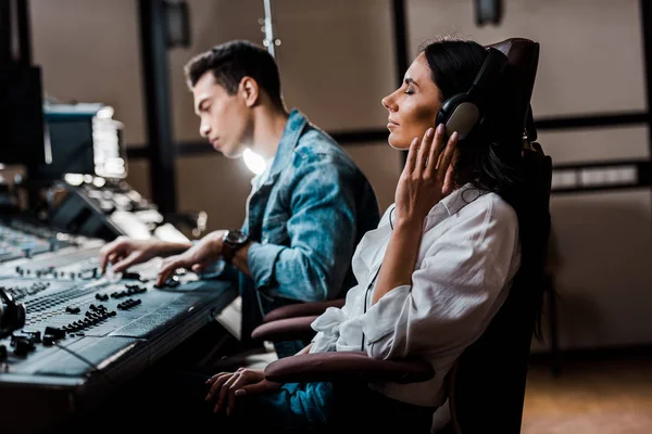 Pretty sound producer in headphones listening music near mixed racial friend working at mixing console — Stock Photo