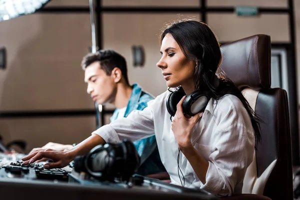 Selective focus of pretty sound producer in earphones working at mixing console near mixed race friend — Stock Photo