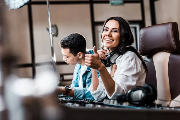 Selective focus of smiling sound produce showing thumb up while working at mixing console near mixed race friend — Stock Photo