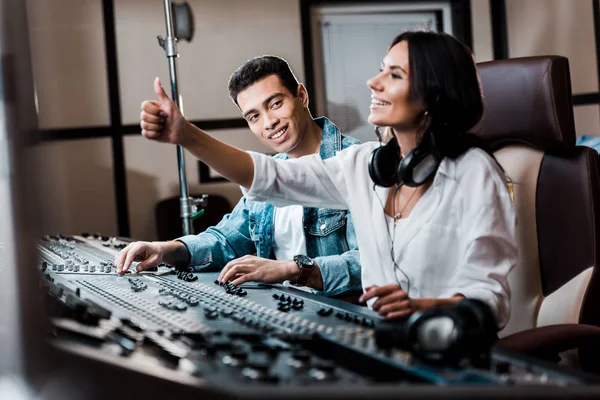 Selective focus of cheerful sound produce showing thumb up while working at mixing console near smiling mixed race friend — Stock Photo