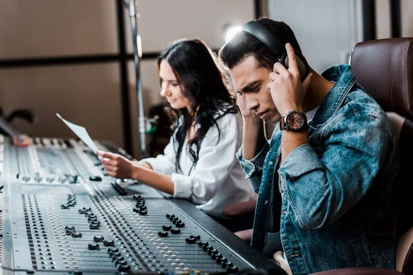 Selective focus of handsome mixed race sound producer in earphones near pretty colleague working at mixing console — Stock Photo