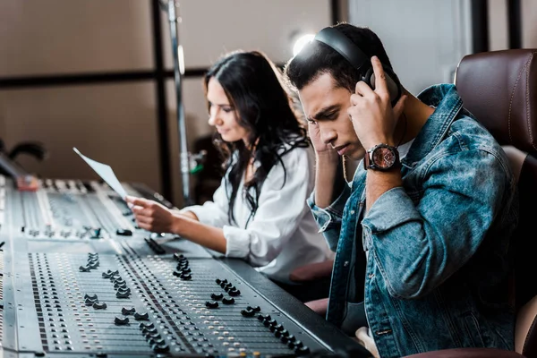 Messa a fuoco selettiva di bel produttore di suoni misti razza in auricolari vicino abbastanza collega che lavora alla console di miscelazione — Foto stock