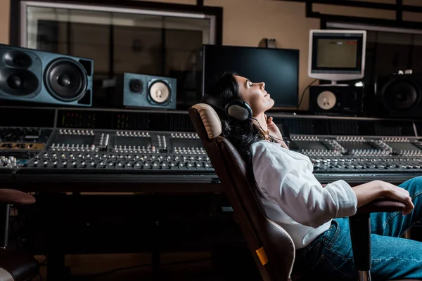 Jolie producteur de son écouter de la musique dans les écouteurs tout en étant assis dans la chaise de bureau dans le studio d'enregistrement — Photo de stock