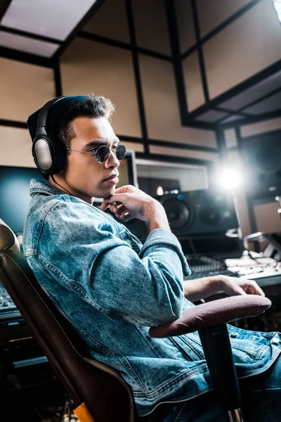 Apuesto serio mezclado raza sonido productor en gafas de sol y auriculares sentado en estudio de grabación - foto de stock