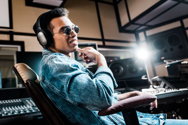 Улыбающийся продюсер смешанных рас в наушниках и солнцезащитных очках, сидящий в студии звукозаписи — стоковое фото