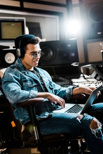 Sonriente productor de sonido de raza mixta en auriculares usando computadora portátil en estudio de grabación - foto de stock