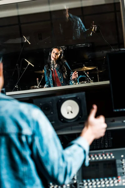 Foyer sélectif du producteur sonore métissé montrant pouce jusqu'à belle chanteuse en studio d'enregistrement — Photo de stock