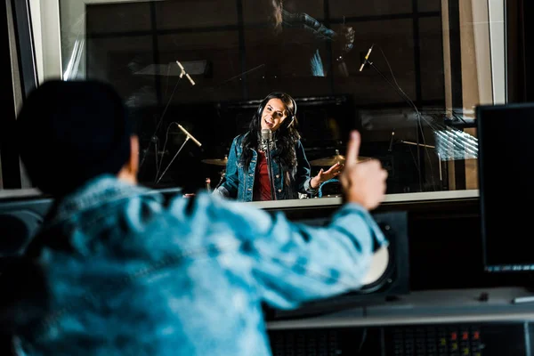 Selective focus of mixed race sound producer showing thumb up to woman singing in recording studio — Stock Photo