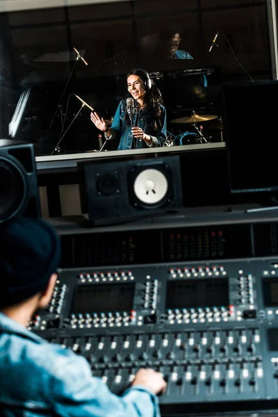 Selective focus of mixed race sound producer working at mixing console while pretty woman singing in recording studio — Stock Photo