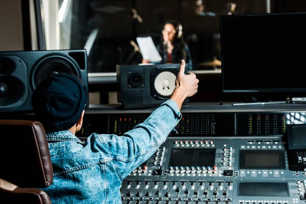 Selective focus of mixed race sound producer showing thumb up to woman singing in recording studio — Stock Photo