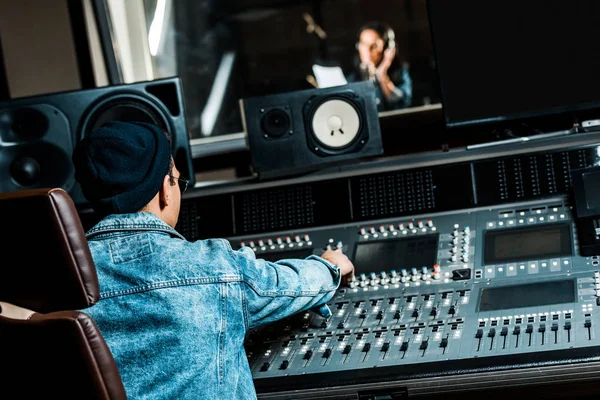 Foco seletivo de produtor de som de raça mista trabalhando no console de mixagem enquanto mulher bonita cantando em estúdio de gravação — Fotografia de Stock
