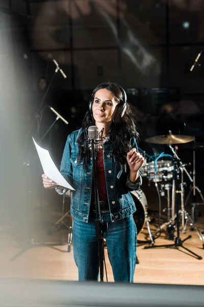 Selective focus of inspired woman in headphones standing near microphone in recording studio — Stock Photo