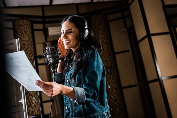 Selective focus of attractive woman holding paper with text and singing in recording studio — Stock Photo